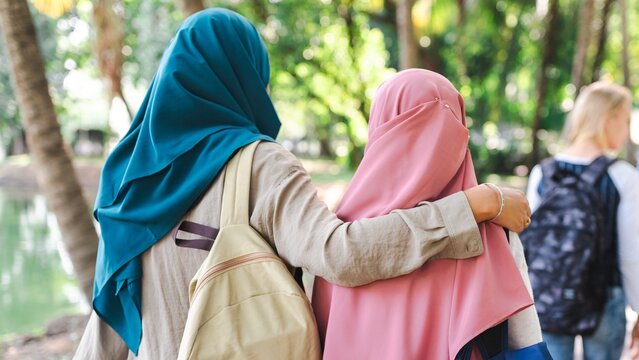 Muslim women in hijabs, walk together in a park. They are embracing, showing friendship and connection. Trees and a pond are visible. International students and muslim girls at school.