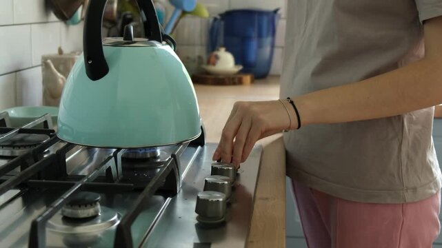 Young blonde woman in a casual outfit lighting the gas stove burner with a lighter to boil water in a turquoise kettle for making tea or coffee in her modern and cozy kitchen