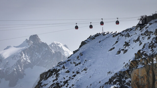 Aerial view of Vallee Blanche SkyWay gondolas suspended over snow-dusted peaks, ascending towards the Aiguille du Midi under a muted sky, Chamonix, Auvergne-Rhone-Alpes, France.
