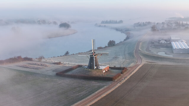Aerial view of a windmill standing proudly amidst a misty landscape, where the calm river reflects the soft morning light, Beesel, Limburg, Netherlands.