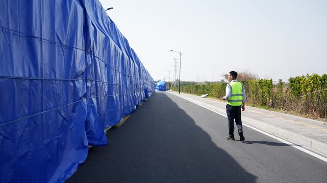 A worker wearing a reflective vest walks along a road lined with blue construction tarps