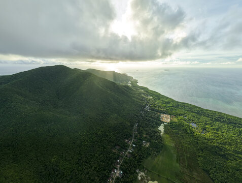 Aerial view of the lush green landscape meets the turquoise ocean under a sky streaked with sunlight and clouds, Karimunjawa, Central Java, Indonesia.