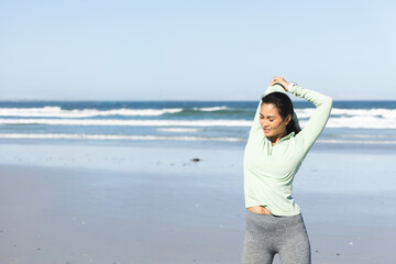 Asian woman stretching on beach with wet sand, wearing green top and grey leggings, copy space