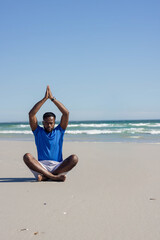 African American man sitting on sandy beach by waterline in blue shirt and wristwatch meditating © wavebreak3