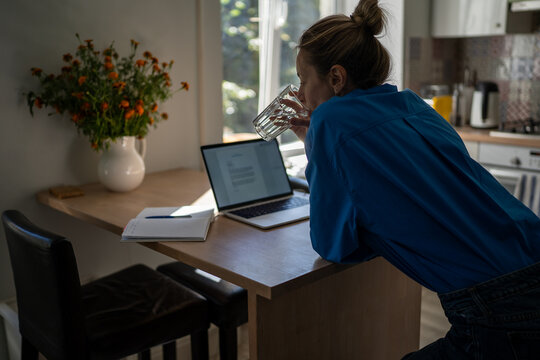 Relaxed mature woman working on laptop in kitchen at home, lady reading notes in notebook and planning workday. Thoughtful female drinking glass of water and thinking, opened computer at table.