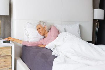 Senior woman lying in bed propping on left elbow reaching for digital alarm clock on nightstand © wavebreak3
