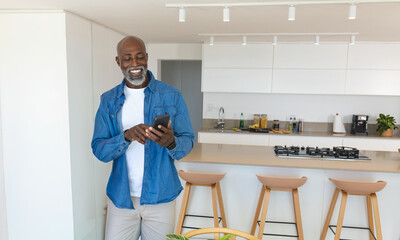 African American man smiling while holding smartphone in modern kitchen near island and stools