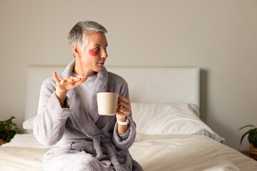Senior woman sitting on bed wearing plush gray robe, holding beige mug, showing red eye patch
