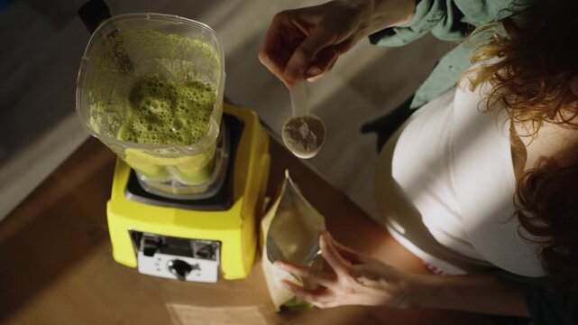 Young pregnant woman focusing on a healthy lifestyle, adding a scoop of protein powder to a fresh green smoothie in a blender for an energy boost and proper nutrition