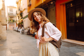 Obraz premium Beautiful tourist wearing hat and carrying backpack strolls through narrow streets of city on sunny day. Young woman enjoys the architecture and the city walk. A weekend, travel concept.