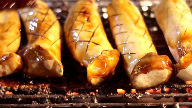 Professional shot of King Oyster mushrooms sizzling on a charcoal grill. The vendor applies a savory glaze that drips over the scored surface creating a delicious vegan street snack.