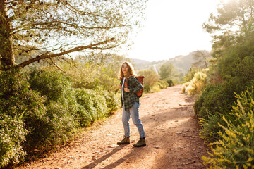 Obraz premium Young woman with hiking backpack walks along mountain path surrounded by green plants. Female hiker enjoys mountain scenery and nature at sunset. Concepts: adventure, relaxation, active lifestyle.