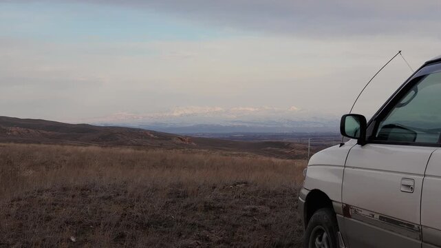Early spring, mountain landscape, low clouds, a white car parked on a hill, looking at the horizon where snow-capped mountains are visible, the wind sways the dry grass