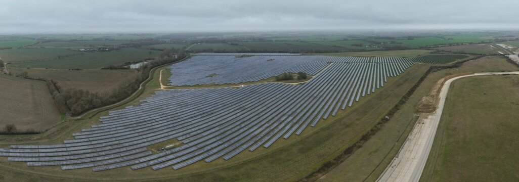 Aerial view of solar panels glinting under a cloudy sky near the former RAF Alconbury, contrasting with the surrounding green fields, Huntingdon, Cambridgeshire, United Kingdom.
