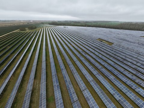 Aerial view of neat rows of solar panels spread across the landscape near the former RAF Alconbury, reflecting the sky's muted tones, Huntingdon, Cambridgeshire, United Kingdom.