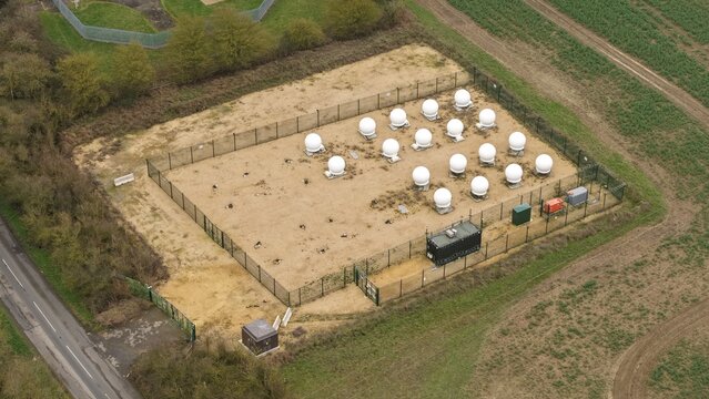 Aerial view of clustered white domes within a fenced perimeter, contrasting against the surrounding muted green and brown fields, Alconbury, Huntingdon, United Kingdom.