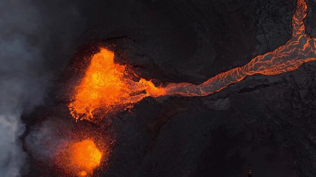 Aerial view of molten lava flowing like a fiery river amid dark volcanic rock, juxtaposed against plumes of grey smoke, Grindavik, Grindavikurbaer, Iceland.