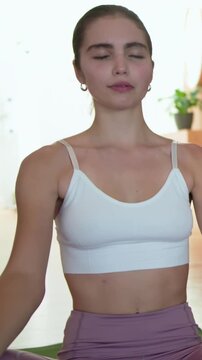 Vertical video: Camera pulling back as woman adjusting white crop top while meditating on green mat