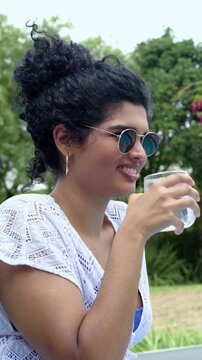 Vertical video: Shifting gaze, woman wearing shades and cover-up lifting glass, sipping in garden