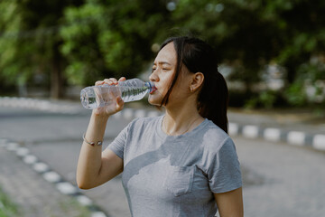 Asian sportswoman making break in jogging, drinking water on bottle in the park. Copy space for text.