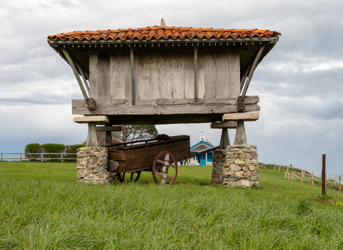 Traditional Spanish horreo granary made of wood and stone on a green field with a wooden cart and a small chapel