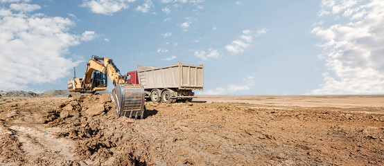 Heavy machinery is active at a construction site with an excavator moving earth while a dump truck waits to collect the dirt under a clear sky © Anoo