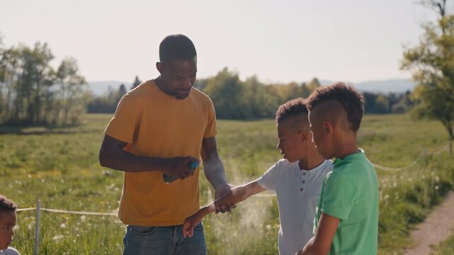 In the countryside, a father looking after his three sons, spraying the older twins with mosquito repellent before they play in nature, medium shot.