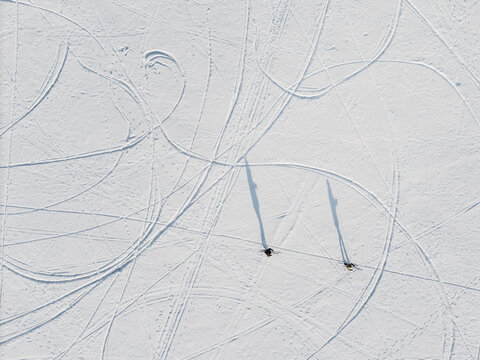Aerial view of two figures casting long shadows across the stark white expanse of frozen Lake Galve, etched with looping ski tracks, Trakai, Lithuania.