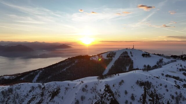 Aerial view of the snow-capped Mottarone mountain at sunset, with the sun reflecting on the lake and the horizon, Mottarone, Piedmont, Italy.