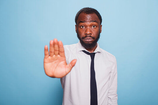 On isolated blue background, serious looking businessman in a white shirt and black tie, makes a hand stop in disapproval. Professional office worker with a grumpy demeanor putting up denial sign.