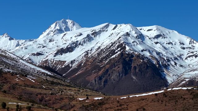 Aerial view of snow-capped mountains contrasting with the clear blue sky, with brown and green foothills below, Campan, Occitanie, France.