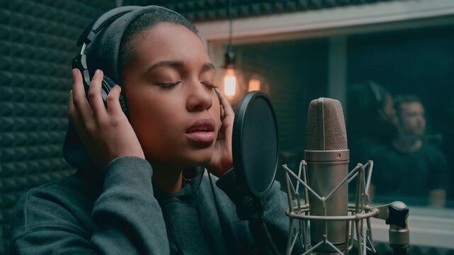 Young African American woman singing passionately into a studio microphone. Female vocalist recording a song in a soundproof booth with headphones. Music production concept