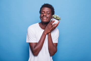 Portrait of african ameriacan individual smiling while holding a flowerpot close to his face. In front of blue background, happy black man carrying a white container with green leaves.