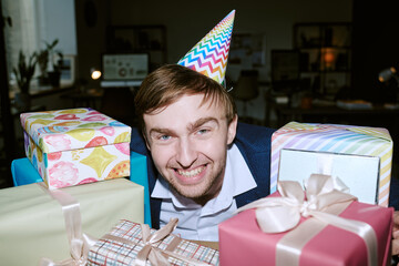 Obraz premium Portrait of young adult Caucasian man smiling and wearing party hat, leaning forward among several wrapped gift boxes, celebrating birthday in indoor setting, looking at camera