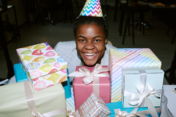 Obraz premium Portrait of Black girl wearing party hat smiling behind stack of wrapped birthday presents, showing joyful expression, sitting at table in indoor celebration setting