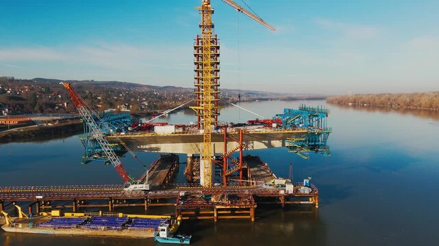 Aerial view of a massive bridge construction with cranes over a wide river, reflecting the clear blue sky, Novi Sad, Vojvodina, Serbia.