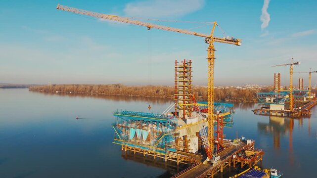 Aerial view of a bridge under construction over a wide river, with cranes and scaffolding, reflecting the clear sky, Novi Sad, Vojvodina, Serbia.