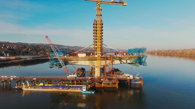 Aerial view of a bridge under construction with yellow cranes reflected in the water, creating a contrast against the clear blue sky, Novi Sad, Vojvodina, Serbia.