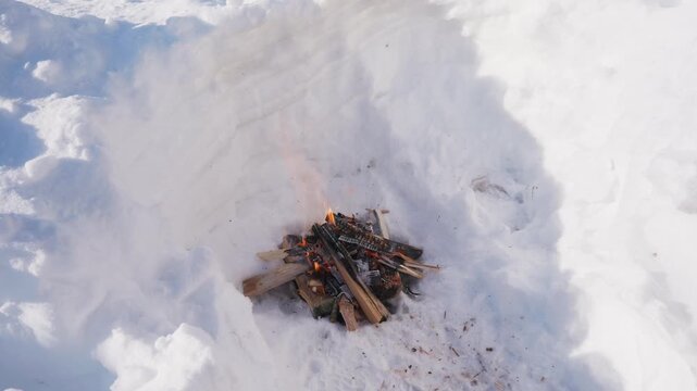 Small campfire made of stacked firewood burning inside a deep snow pit in winter. Flames flicker between logs while the fire burns outdoors in cold snowy conditions. High quality 4k footage