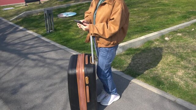 Woman holds a mobile phone in her hands and checks the check-in time through the app. Woman with a suitcase calls a taxi to the airport