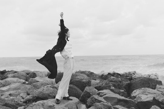 Woman balances on jagged rocks beside the sea in a monochrome scene, wind catching fabric, calm strength, open horizon, dynamic gesture of resilience and freedom.