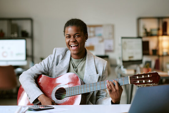 Portrait of young adult Black woman smiling while playing acoustic guitar at desk in modern office setting, showing joyful expression and engaging with musical instrument indoors