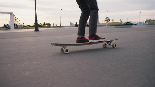Man's legs and feet skillfully riding a longboard on an asphalt surface in a contemporary city park, with people and architecture visible in the background during a beautiful sunset