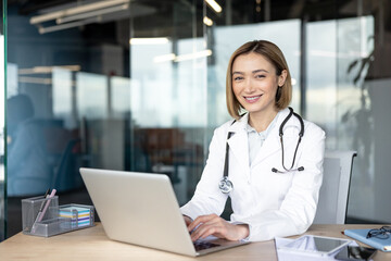 Female doctor working on a laptop, smiling at the camera, wearing a lab coat and stethoscope in a modern office, representing healthcare innovation and professional medical consultation