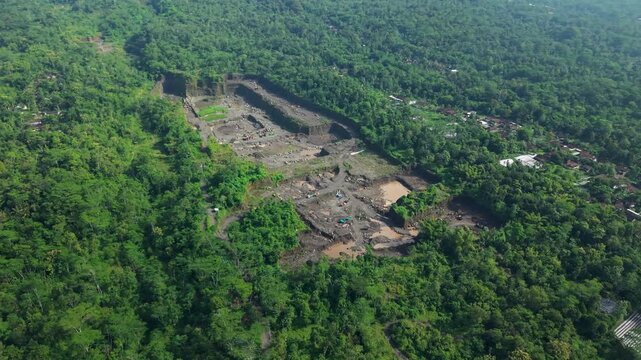 Aerial view of a quarry cutting into the earth, surrounded by dense green trees, creating a stark contrast between the natural and industrial at Kali Woro, Klaten, Jawa Tengah, Indonesia.