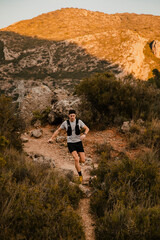 Obraz premium Young trail runner running along a steep mountain path surrounded by rugged scenery and the warm light of the sunset. Outdoor endurance training and adventure sport concept.