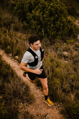 Fototapeta premium Athletic trail runner running along a winding mountain trail surrounded by wild vegetation and rugged terrain. Outdoor endurance sport and adventure lifestyle in nature.