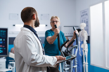 Obraz premium At physiotherapy clinic, doctor with tablet, addresses elderly patient who is doing gymnastics for recovery. While speaking with physician, old woman exercises on stationary bicycle.