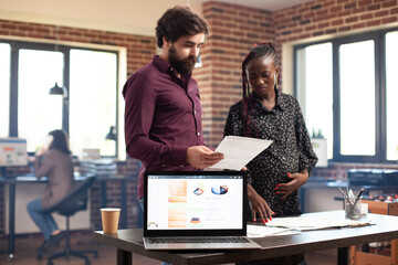 Fototapeta premium Black expectant mother discussing with male colleague as financial chart is shown on laptop screen. Digital device displaying company revenues as white man brainstorms with pregnant lady nearby.