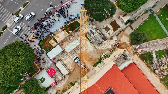 Aerial view of a vibrant construction site featuring a towering yellow crane juxtaposed against the red roof, and green trees, Chania, Chania, Greece.